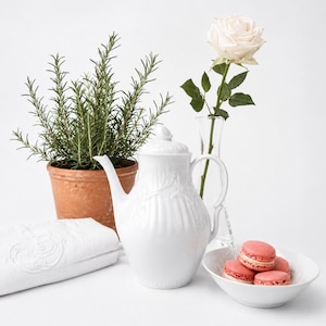 Peut inclure: Une théière en porcelaine blanche avec un corps et un couvercle côtelés, un pot en terre cuite contenant un romarin vert, une rose blanche dans un vase en verre, un tissu brodé blanc et un bol rempli de macarons roses sur une surface blanche.