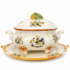Peut inclure: Une soupière en céramique blanche avec son couvercle et son plat de service assortis. La soupière et l'assiette sont décorées de motifs floraux et d'oiseaux jaunes et bleus. La soupière a deux anses et repose sur trois petits pieds.