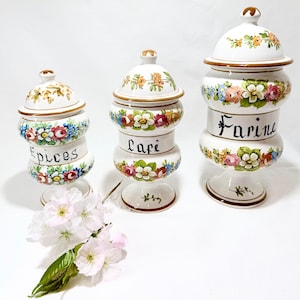Puede incluir: Tres botes de cerámica blanca para especias con tapas, adornados con coloridos diseños florales y ribetes dorados. Los botes están etiquetados como "Epices", "Cafe" y "Farine". Una ramita de flores rosas descansa frente a los botes.