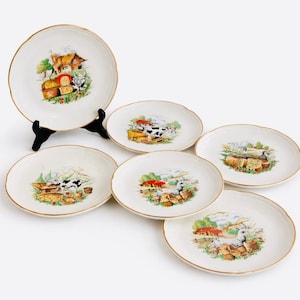 May include: A set of six white ceramic plates with a gold rim. Each plate features a colorful illustration of a farm scene with a house, cow, and cheese. One plate is displayed on a black stand.
