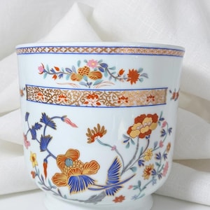 Könnte beinhalten: Weiße Keramiktasse mit floralem Design in Orange, Blau und Gelb. Die Tasse hat ein dekoratives Band mit goldenen und braunen Akzenten. Das florale Muster umfasst stilisierte Blumen und Blätter. Die Tasse hat einen Sockel.