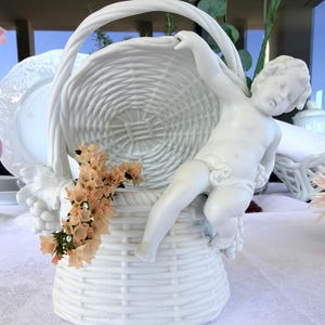 Peut inclure: Un panier en porcelaine blanche avec une texture tressée et une anse haute arquée. Une figurine sculptée de chérubin est fixée sur le côté du panier, et un bouquet de petites fleurs séchées de couleur pêche pâle est inséré à l'avant.