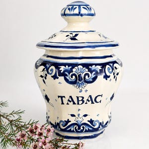 Peut inclure: Un pot en céramique blanc avec un couvercle, orné de motifs floraux bleus et du mot « TABAC » en lettres grasses. Le pot a une forme bombée et est décoré de motifs bleus complexes. Une branche de fleurs roses repose près du pot.