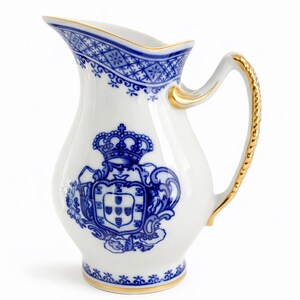 May include: White porcelain pitcher with blue and gold accents. The pitcher features a blue crest with a crown and intricate patterns. The handle and rim are trimmed with gold. A decorative piece for serving.
