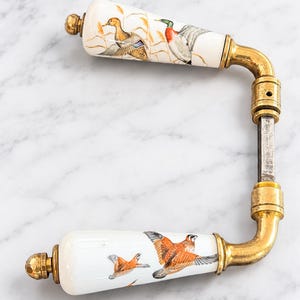 Pode incluir: Um conjunto de maçanetas para porta com dois puxadores de cerâmica branca decorados com ilustrações detalhadas de patos e aves de caça. Os puxadores estão ligados por um eixo central de metal e possuem ferragens em tom de latão envelhecido.