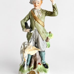 Könnte beinhalten: Porzellanfigur eines Jägers in einem grünen Mantel und grauem Hut, der ein Gewehr hält und mit einem Hund steht. Der Jäger hat eine goldene Schärpe und steht auf einem Baumstumpf mit grünem Laub. Der Sockel ist mit goldenen Akzenten verziert.