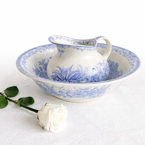 Peut inclure: Un pichet et un bassin en céramique blanche avec des motifs floraux et paysagers bleus. Le pichet est posé dans le bassin, et une rose blanche avec des feuilles vertes est posée sur le côté. L'ensemble a une esthétique vintage.