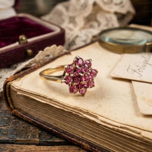 May include: A gold ring with a cluster of small, round, red gemstones arranged in a floral pattern. The ring sits on an open antique book with aged pages. A vintage jewelry box and magnifying glass are in the background.