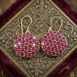 May include: A pair of circular drop earrings featuring a cluster of small, round, vibrant pinkish-red gemstones set in gold metal. Each earring is suspended from a curved gold wire hook, showcasing a dense, pavé-style arrangement of stones.
