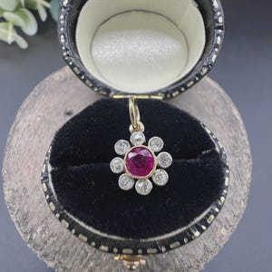 Puede incluir: Un colgante de oro con un rubí central de color rosa intenso de talla redonda, rodeado por un halo de ocho pequeños diamantes blancos de talla redonda dispuestos en forma de flor, presentado en un joyero circular forrado de terciopelo negro.