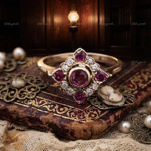 May include: A gold ring featuring a central round-cut deep pink gemstone surrounded by a cluster of smaller white stones and four larger round-cut pink stones arranged in a diamond-shaped setting on a polished gold band.