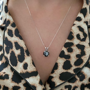 May include: A round, faceted black gemstone pendant set in a silver-toned four-prong metal setting, suspended from a delicate, thin silver-toned chain. The pendant rests against a garment featuring a tan and black leopard print pattern.