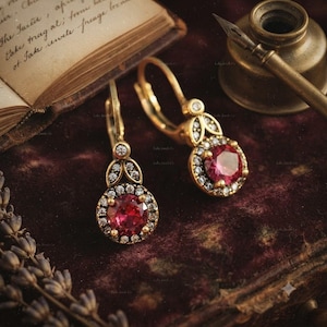 May include: A pair of gold-toned drop earrings featuring a central round-cut red gemstone surrounded by a halo of small clear stones. The earrings have a lever-back closure and a decorative leaf-shaped accent set with tiny clear stones above the center.