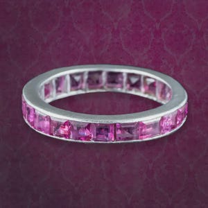 Op de afbeelding: Een zilverkleurige eternity-ring met een doorlopende rij vierkant geslepen roze edelstenen gezet in een gepolijst metalen frame. De ring heeft een uniform ontwerp waarbij elke steen stevig op zijn plaats zit voor een levendige uitstraling.