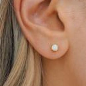 May include: A small, round stud earring featuring a central white opal gemstone set in a polished gold-tone metal base. The earring is positioned centrally on the earlobe, showcasing a smooth, iridescent surface that reflects light with subtle flashes.