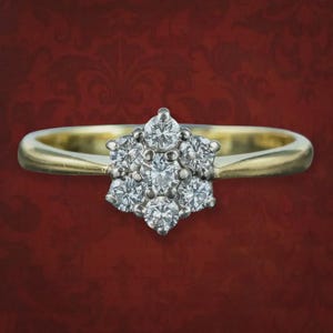 May include: A gold ring featuring a cluster of seven round, clear, faceted stones arranged in a floral pattern. The central stone is surrounded by six smaller stones, all set in a metal mount that transitions into a smooth, polished gold band.