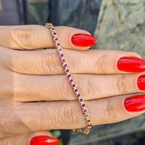 May include: A gold tennis bracelet featuring a continuous row of small, round, bezel-set red rubies. The bracelet is composed of individual circular metal settings, each housing a single vibrant red gemstone, linked together to form a flexible chain.
