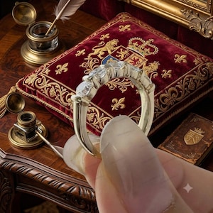 May include: A silver-toned ring featuring five round, clear, faceted stones set in a row along the top half of the band. The band has a smooth, polished finish and tapers slightly toward the bottom, showcasing a classic, symmetrical design.