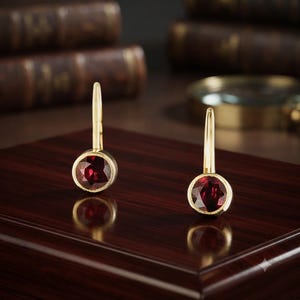 Puede incluir: Un par de pendientes de oro con cierre de palanca, cada uno con una piedra preciosa redonda de color rojo intenso facetada y engastada en un bisel de oro pulido. Los pendientes tienen un diseño clásico de gota con un mecanismo de alambre curvo.