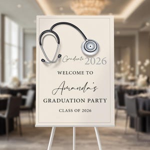 May include: A graduation party welcome sign featuring a stethoscope graphic. The sign reads "Graduate 2026" and "Welcome to Amanda's Graduation Party Class of 2026" in elegant script and sans-serif fonts. The sign is displayed on a white easel.