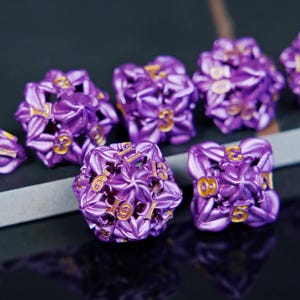 May include: A collection of eight purple polyhedral dice with gold numbering. The dice have a floral design and are arranged on a dark surface. The dice are used for tabletop role-playing games.