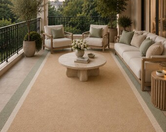 May include: A large rectangular area rug featuring a solid tan centre, a wide cream-coloured inner border, and a sage green outer border. The rug is positioned on a tiled balcony floor beneath a sofa, two armchairs, and a round stone coffee table.