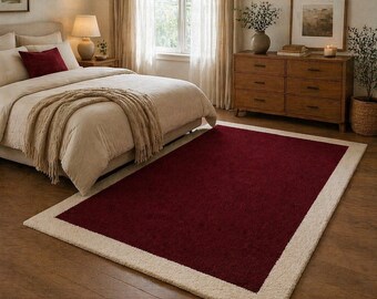 May include: A rectangular rug with a burgundy centre and a cream-coloured border. The rug is placed on a hardwood floor in a bedroom setting, complementing the neutral tones of the bed and furniture.