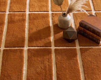 May include: A textured area rug featuring a repeating pattern of burnt orange rectangular blocks separated by thin white lines. A ceramic vase with dried foliage, a dark geometric object, and a stack of three leather-bound books sit on top.