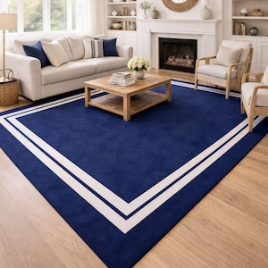 Puede incluir: Una alfombra rectangular grande que presenta un campo azul marino profundo con un borde prominente de doble línea blanca que sigue el perímetro. La alfombra está colocada sobre un suelo de madera clara en una sala con muebles neutros.