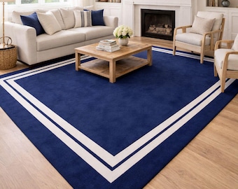 May include: A large rectangular area rug featuring a deep navy blue field with a prominent white double-line border that follows the perimeter. The rug is positioned on a light-toned hardwood floor in a living room setting with neutral furniture.