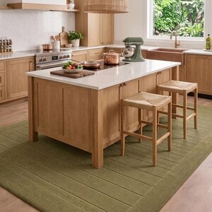 Puede incluir: Una isla de cocina con encimera blanca, armarios de madera clara y dos taburetes de madera con asientos trenzados. Una batidora verde, utensilios de cobre y un bol de verduras est&aacute;n sobre la isla. Una gran alfombra verde oliva cubre el suelo.