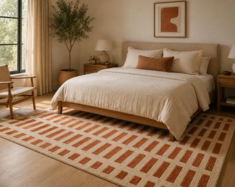 May include: A bedroom scene with a bed covered in beige bedding and a rust-coloured cushion. A patterned rug with rust-coloured rectangular blocks is on the floor. An abstract artwork hangs above the bed.