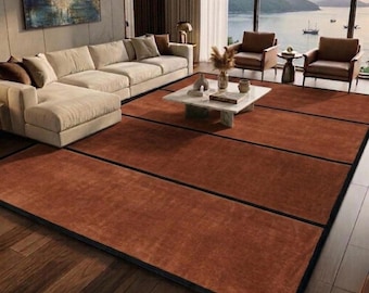 May include: A large rectangular area rug featuring a solid terracotta orange surface divided by thin black grid lines. The rug is positioned on a dark wooden floor in a living room setting, extending from the foreground toward a white sofa and chairs.