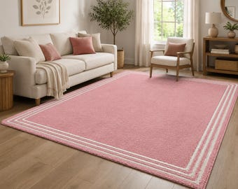 May include: A large, rectangular pink rug with a white border and two white stripes. The rug is placed on a wooden floor in a living room setting, with a sofa, chair, and side table visible in the background.