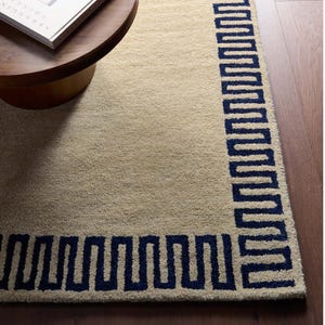 Puede incluir: Una alfombra rectangular con un centro beige sólido y un borde azul marino oscuro con un patrón geométrico de greca repetitivo. La alfombra tiene una superficie de pelo texturizado y copetudo, colocada sobre un suelo de madera marrón oscuro.