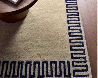 May include: A rectangular area rug featuring a solid beige centre and a dark navy blue border with a repeating geometric Greek key pattern. The rug has a textured, tufted pile surface and is positioned on a dark brown wooden floor.