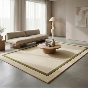 Puede incluir: Un salón de tonos neutros con una alfombra beige con borde verde. Un sofá bajo beige se encuentra junto a una ventana con cortinas transparentes. Una mesa de centro redonda de madera exhibe objetos decorativos. Una lámpara escultural y una obra de arte completan el espacio.