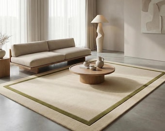 May include: A modern living room with a beige rug and a green border. A beige sofa is positioned near a window with sheer curtains. A round wooden coffee table displays decorative items. A tall, sculptural lamp and artwork add to the room's design.