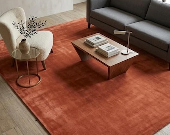 May include: A large, rectangular area rug in a solid, deep terracotta orange colour. The rug features a short, dense pile texture and covers a significant portion of the light-coloured wooden floor, serving as a foundation for the surrounding furniture.