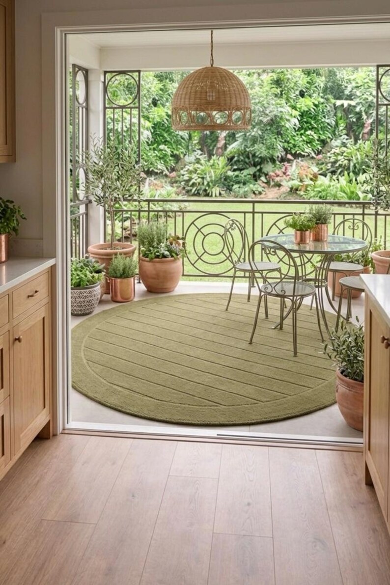 Puede incluir: Escena de patio al aire libre con una alfombra redonda verde oliva con dise&ntilde;o texturizado. Una mesa con tapa de cristal y sillas est&aacute;n dispuestas en el patio, rodeadas de plantas en macetas y una l&aacute;mpara colgante tejida.
