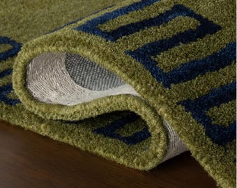 May include: A thick, tufted area rug featuring a deep olive green base colour with a bold, dark navy blue geometric pattern. The rug is folded over to reveal a light-coloured, woven backing material and the dense, textured pile of the fabric surface.