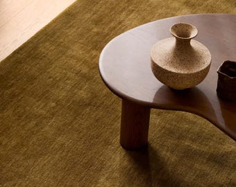 May include: A dark olive green area rug with a textured surface, featuring a dark brown wooden coffee table with a curved edge and a speckled beige ceramic vase with a narrow neck and a wide base placed on top of the table surface.