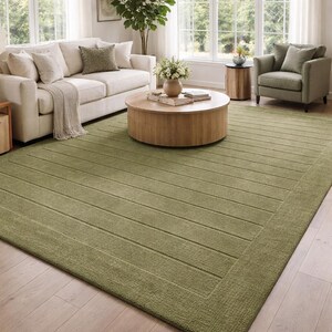 Hand tufted sage green wool area rug with subtle raised linear texture displayed in a bright modern living room with cream sofa, natural wood coffee table, green accent chair, large windows and light wood flooring.