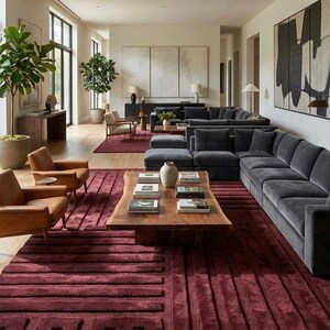Puede incluir: Una sala de estar grande con una alfombra color burdeos profundo con un patrón geométrico en relieve, un sofá seccional gris oscuro, dos sillones de cuero color canela y una mesa de centro de madera natural con borde irregular.