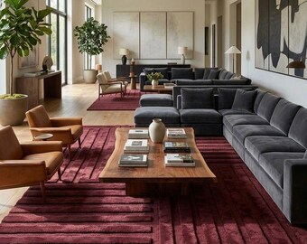 May include: A large living room featuring a deep burgundy area rug with a raised geometric pattern, a dark grey sectional sofa, two tan leather armchairs, and a natural wood coffee table with a live edge, all set on light wood flooring.
