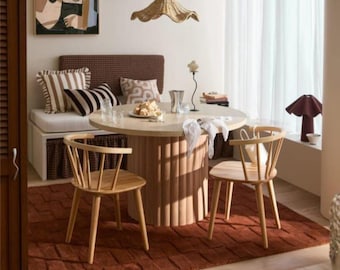 May include: A dining area featuring a round light-coloured table with a fluted base, two light wood spindle chairs, and a bench with brown patterned cushions. A woven pendant light hangs above, and a textured rust-coloured rug covers the floor.