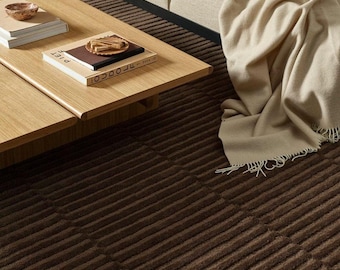May include: A dark brown area rug with a textured, ribbed, or striped pattern covering the floor. The rug features a consistent linear design across its surface, providing a deep, solid colour foundation beneath a light wood coffee table and sofa.