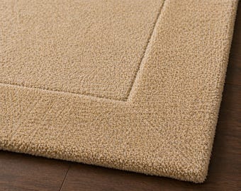 May include: Close-up of a beige rectangular rug with a textured surface and a defined border. The rug is made of a soft, plush material, and the border creates a framed effect. The rug is placed on a dark wood floor.