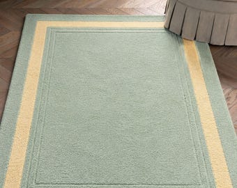 May include: A rectangular rug in a light green hue, framed by a yellow border. The rug is placed on a wooden floor, with a chair and curtains in the background. The rug has a soft, textured appearance.