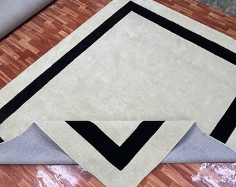 May include: A cream-coloured rectangular rug with a thick black border. A section of the rug is folded over, revealing a black chevron design on the corner. The rug is on a wooden floor.
